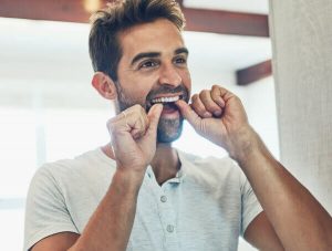 Halitosis Wimbledon - A man is practicing good oral hygiene by flossing his teeth in a bright, modern bathroom. Ridgway Dental, a dental clinic in Wimbledon, offers treatments and advice to combat halitosis and other oral health issues. The image emphasizes the importance of daily dental care routines in preventing bad breath and maintaining overall oral health. The man appears focused and content, illustrating the positive impact of effective dental habits. Ridgway Dental provides comprehensive care and guidance to help patients in Wimbledon achieve fresh breath and healthy teeth.