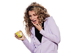 Knocked-Out Tooth Wimbledon - A woman in a light purple hoodie grimaces in pain, holding her jaw with one hand and an apple in the other, suggesting she may have experienced a knocked-out tooth while eating. The white background focuses attention on her discomfort. Ridgway Dental, a dental clinic in Wimbledon, provides specialized care for dental emergencies such as knocked-out teeth. This image highlights the importance of seeking immediate professional dental treatment to preserve the tooth and alleviate pain. The clinic's expertise ensures that patients receive the necessary care to manage and treat such dental traumas effectively.