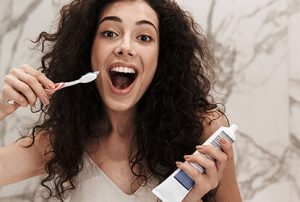 Prevent Dental Emergencies with Proper Brushing Wimbledon - A woman with curly hair is enthusiastically brushing her teeth in a bright, modern bathroom. She holds a toothbrush in one hand and a tube of toothpaste in the other, smiling widely at the camera. The lively and positive image emphasizes the importance of good oral hygiene. Ridgway Dental, a dental clinic in Wimbledon promotes such preventive measures to avoid dental emergencies. Regular brushing, as demonstrated by the woman, is essential for maintaining healthy teeth and gums. The clean and stylish bathroom setting reflects the quality care and professional environment provided by Ridgway Dental in Wimbledon.