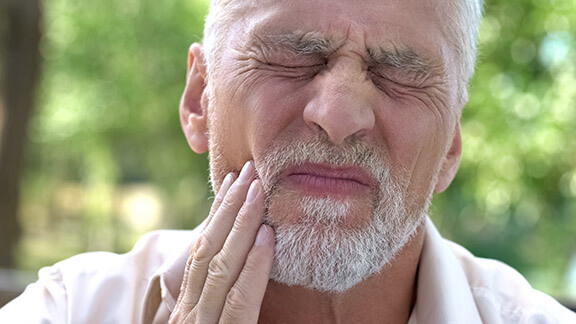 Emergency dentist Wimbledon - An elderly man grimaces in pain, clutching his jaw, indicating severe dental discomfort, possibly a toothache or dental emergency. The background suggests an outdoor setting with blurred greenery, emphasizing the urgency of seeking immediate dental care. This image underscores the need for prompt attention from an emergency dentist in Wimbledon. Ridgway Dental, a dental clinic in Wimbledon, provides swift and effective relief for such dental emergencies, ensuring patients receive the necessary care to alleviate pain and address urgent dental issues.